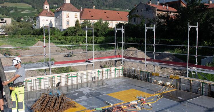 Ein Bagger gräbt in einem großen Erdloch auf der Baustelle des Hofburg Gartens; im Hintergrund Sträucher, die Türme des Doms und bewaldete Berge.
