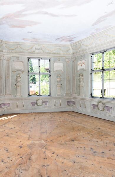 Historischer Raum mit hellen Wandmalereien, großen Fenstern und verzierter Decke.